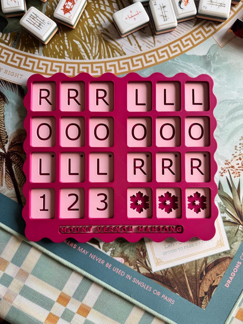 May include: A bright pink, scalloped-edge Mahjong tile rack featuring a grid of sixteen square cutouts. The rack displays the letters R, L, O, and the numbers 1, 2, 3, along with flower icons. The text MOUNT VERNON MAHJONG is embossed at the bottom.
