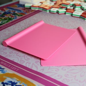 May include: Two rectangular, solid pink plastic mahjong tile racks with a smooth, flat surface and a rounded, cylindrical edge along the top. These lightweight game accessories are designed to hold tiles upright during play, featuring a simple, clean design.