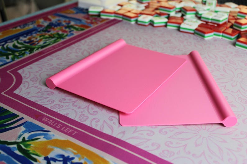 May include: Two rectangular, solid pink plastic mahjong tile racks with a smooth, flat surface and a rounded, cylindrical edge along the top. These lightweight game accessories are designed to hold tiles upright during play, featuring a simple, clean design.