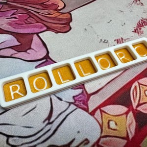 May include: A rectangular white plastic frame containing six individual square compartments, each filled with a shimmering golden-yellow resin. The white letters R, O, L, L, O, R are embedded within the resin, spelling out the word ROLLOR.