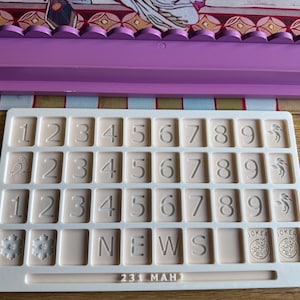 May include: A beige number and letter tray with embossed numbers, letters, and symbols. The tray includes the word "NEWS", the numbers "231", and the word "MAH". The tray also features snowflake, bird, and joker symbols.