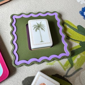 May include: A square, olive green coaster with a wavy lavender border holds a white rectangular card featuring a palm tree illustration. Other cards with animal illustrations are also visible.