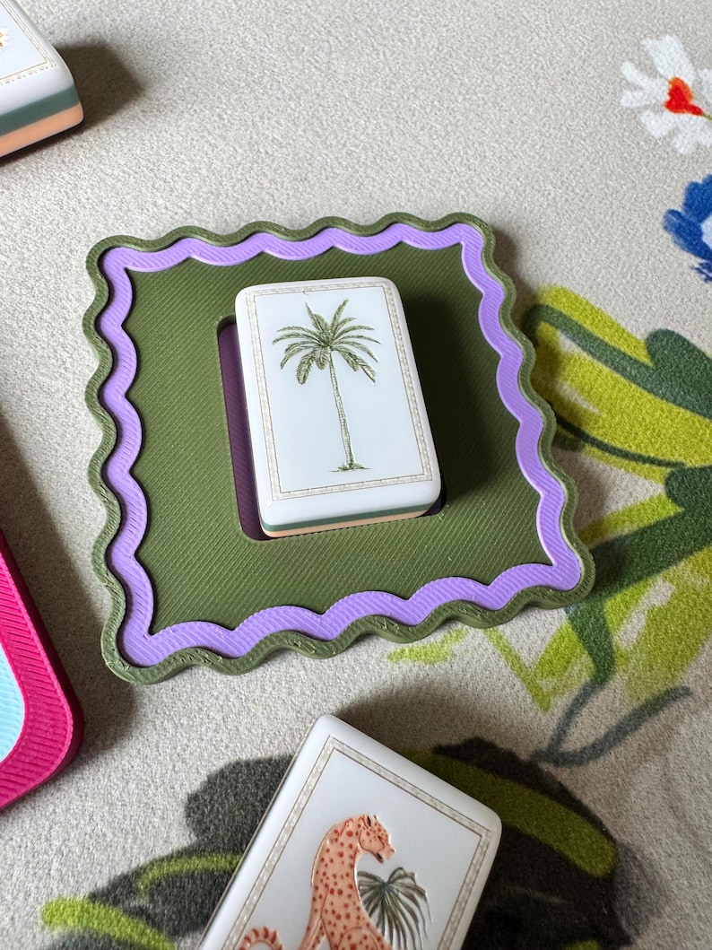 May include: A square, olive green coaster with a wavy lavender border holds a white rectangular card featuring a palm tree illustration. Other cards with animal illustrations are also visible.
