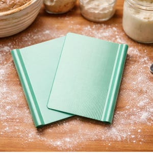 May include: Two mint green baking mats with ridged edges are displayed on a wooden surface dusted with flour. The mats are rectangular and appear to be made of silicone. Baking supplies are in the background.