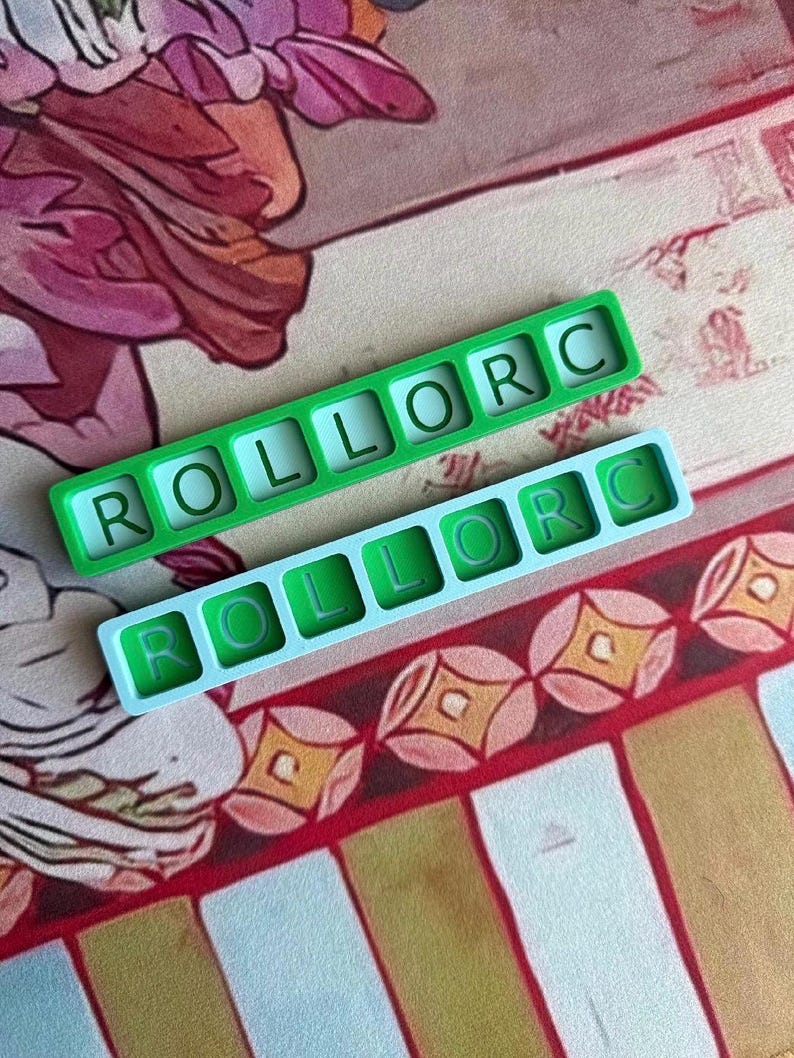 May include: Two rectangular silicone molds, one in bright green and one in light blue, each featuring six square cavities arranged in a single row. Each cavity contains a raised letter, spelling out the word ROLLORC in a clear, sans-serif font.