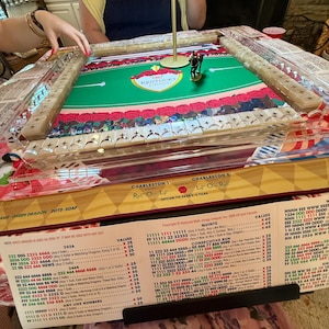 Puede incluir: Un juego de Mah Jongg con una superficie de juego de fieltro verde con el logotipo del Kentucky Derby, rodeada de soportes de azulejos de acr&iacute;lico transparente. Una peque&ntilde;a figura de caballo se encuentra en el centro de la mesa.