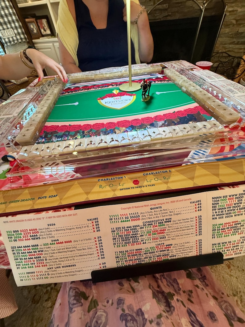 Puede incluir: Un juego de Mah Jongg con una superficie de juego de fieltro verde con el logotipo del Kentucky Derby, rodeada de soportes de azulejos de acr&iacute;lico transparente. Una peque&ntilde;a figura de caballo se encuentra en el centro de la mesa.