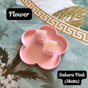May include: A matte sakura pink flower-shaped tray containing two translucent pink dice with gold pips. The tray features the text "Flower" and "Sakura Pink (Matte)" in black boxes. The dice display gold dots on their faces.