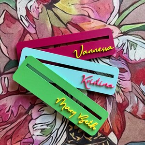 May include: Three rectangular bookmarks featuring horizontal slots and raised lettering. The top bookmark is magenta with yellow text reading Vannessa. The middle is light blue with pink text reading Kristina. The bottom is green with yellow text reading Mary Beth.