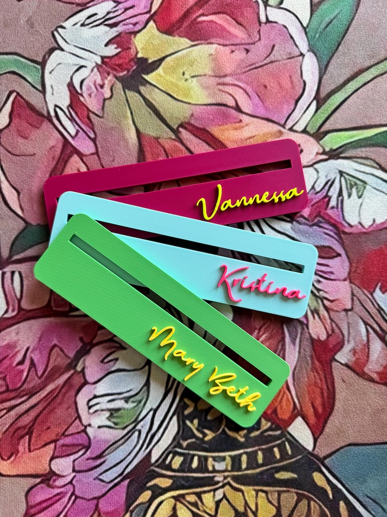 May include: Three rectangular bookmarks featuring horizontal slots and raised lettering. The top bookmark is magenta with yellow text reading Vannessa. The middle is light blue with pink text reading Kristina. The bottom is green with yellow text reading Mary Beth.