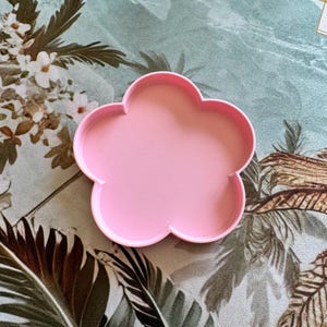 May include: A light pink cookie cutter shaped like a five-petaled flower. The object features a smooth, raised rim and a flat base, designed with a simple, symmetrical outline that forms the distinct silhouette of a blossom with rounded petals.