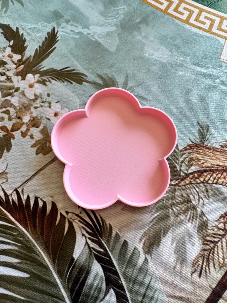 May include: A light pink cookie cutter shaped like a five-petaled flower. The object features a smooth, raised rim and a flat base, designed with a simple, symmetrical outline that forms the distinct silhouette of a blossom with rounded petals.