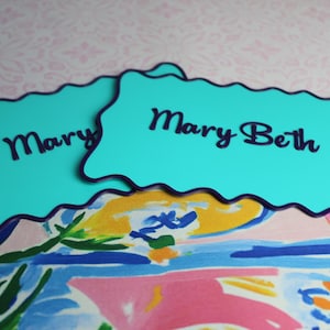 May include: Two teal name cards with a dark blue border and the names "Mary" and "Mary Beth" in dark blue script. The cards are on a colorful floral and patterned background.