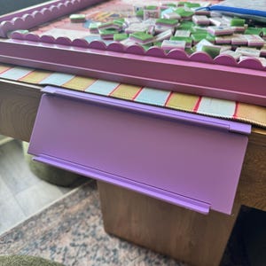 Puede incluir: Un soporte para fichas de pl&aacute;stico de color morado claro con forma rectangular y un borde frontal plano, dise&ntilde;ado para colgarse del borde de una mesa. El soporte presenta una superficie lisa y un dise&ntilde;o sencillo y funcional.