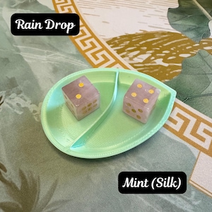 May include: A light mint green, leaf-shaped divided tray containing two pale pink dice with gold-colored pips. The tray is labeled Rain Drop and Mint (Silk). The dice are translucent with rounded corners and gold circular markings on each face.