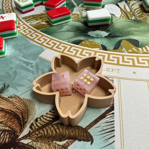 May include: A light brown, flower-shaped tray holds two translucent, pale pink dice with gold-colored pips. The tray is positioned on a surface featuring a green and white botanical pattern, surrounded by several scattered red, white, and green mahjong tiles.