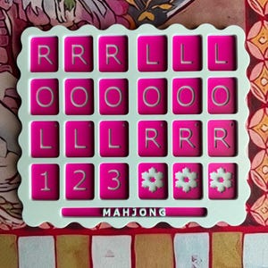 May include: A square, scalloped-edge white game board featuring a grid of twenty-four magenta tiles. The tiles display the letters R, L, and O, the numbers 1, 2, and 3, and snowflake icons. The word MAHJONG is printed in white at the bottom.