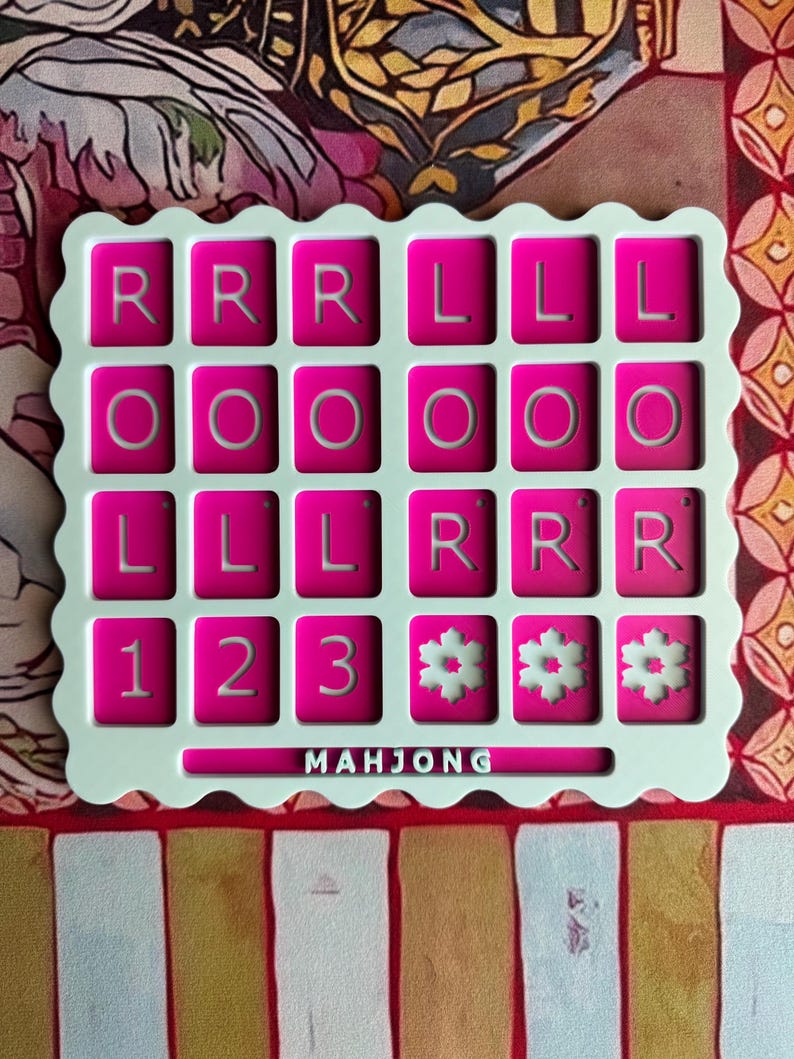 May include: A square, scalloped-edge white game board featuring a grid of twenty-four magenta tiles. The tiles display the letters R, L, and O, the numbers 1, 2, and 3, and snowflake icons. The word MAHJONG is printed in white at the bottom.