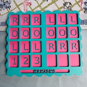 May include: A rectangular teal plastic tray with a scalloped edge featuring a grid of pink recessed squares. The squares contain the letters R, L, O, and the numbers 1, 2, 3 in teal. The word MAHJONG is embossed in teal at the bottom center.