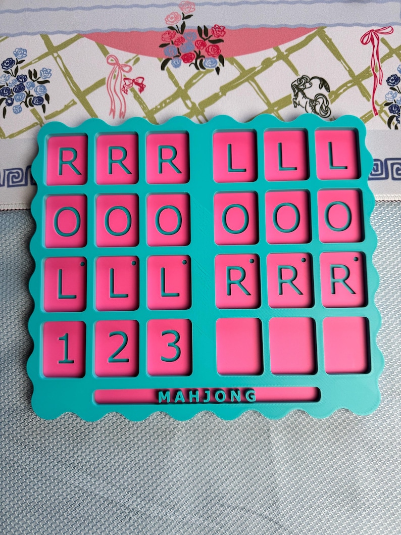 May include: A rectangular teal plastic tray with a scalloped edge featuring a grid of pink recessed squares. The squares contain the letters R, L, O, and the numbers 1, 2, 3 in teal. The word MAHJONG is embossed in teal at the bottom center.
