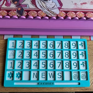 May include: A teal Mahjong tile holder with white tiles featuring numbers, letters, and symbols. A lavender tile rack sits above it. The word "NEWS" is visible on the tiles. The game is on a wooden surface.