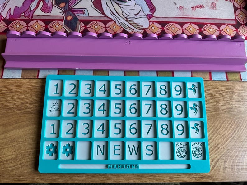 May include: A teal Mahjong tile holder with white tiles featuring numbers, letters, and symbols. A lavender tile rack sits above it. The word "NEWS" is visible on the tiles. The game is on a wooden surface.