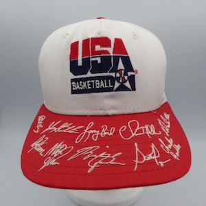 Puede incluir: Gorra de béisbol blanca y roja con el logo "USA Basketball" en rojo y azul marino. La visera roja está adornada con firmas blancas. La gorra tiene un ajuste clásico y estructurado.