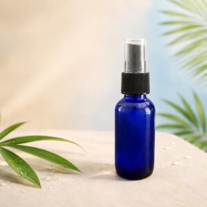 May include: A small, cylindrical cobalt blue glass bottle featuring a black plastic spray nozzle and a clear plastic cap. The bottle has a smooth, reflective surface and is designed for dispensing liquids in a fine mist through the pump mechanism.