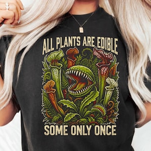 May include: A black t-shirt featuring a graphic illustration of various carnivorous plants, including a large Venus flytrap with sharp teeth. The text "ALL PLANTS ARE EDIBLE" is printed above the plants, and "SOME ONLY ONCE" is printed below.