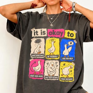 Puede incluir: Una camiseta gris carbón con un estampado gráfico que incluye el texto "it is okay to" sobre seis paneles rectangulares. Cada panel muestra un ganso blanco con los textos: "HAVE HARD DAYS," "BE YOURSELF," "ASK FOR HELP," "START OVER AGAIN," "FEEL YOUR FEELINGS," y "MAKE MISTAKES."