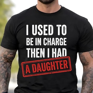 Może przedstawiać: Czarny t-shirt z białym napisem: "I USED TO BE IN CHARGE THEN I HAD A DAUGHTER". Tekst "A DAUGHTER" znajduje się w czerwonym prostokącie. Koszulka ma krótkie rękawy.