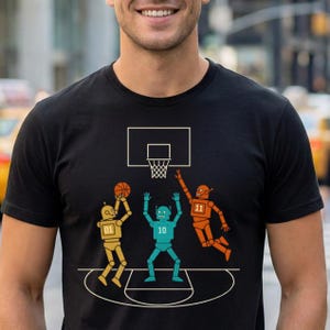 May include: Black t-shirt with a basketball-themed graphic. The design shows three robots playing basketball, with one shooting, one jumping, and another ready to catch. The robots are coloured gold, teal, and orange.