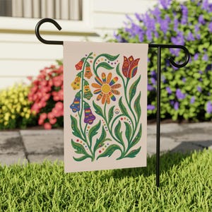 May include: A garden flag featuring a colorful floral design with a rainbow daisy, bell-shaped flowers, and a tulip. The flag has a beige background with green leaves and peace symbols. The flag is displayed on a black metal stand in a grassy yard.