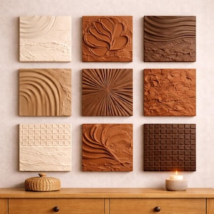 Puede incluir: Nueve paneles cuadrados de arte mural texturizado en tonos beige, marrón y chocolate. Los paneles presentan varios diseños, incluyendo una flor, olas y un patrón de cuadrícula. Una pequeña cesta tejida y una vela encendida en un soporte de hormigón están sobre una superficie de madera.