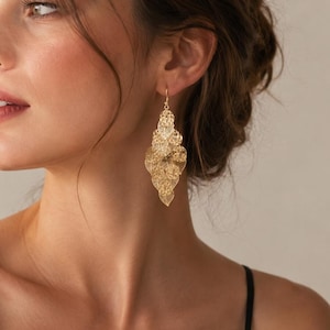 Peut inclure: Une boucle d'oreille pendante dorée présentant un design en feuilles superposées en filigrane avec une texture maillée délicate et complexe. La boucle d'oreille est suspendue à un crochet fin, affichant un agencement étagé de plusieurs éléments en forme de feuille avec une finition métallique brillante.