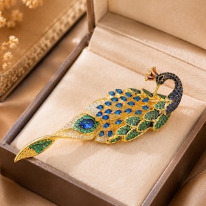 Puede incluir: Un broche de pavo real en tono dorado con un diseño detallado incrustado con pequeños cristales azules, verdes y transparentes. La cabeza del pavo real es azul oscuro, el cuerpo muestra plumaje azul y verde, y la larga cola tiene piedras claras.