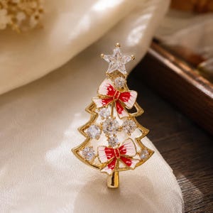 May include: A gold-toned Christmas tree brooch featuring a clear star-shaped crystal at the top, two red and white enamel bows, and multiple round clear crystals embedded throughout the tree structure, all set within a metallic gold frame.