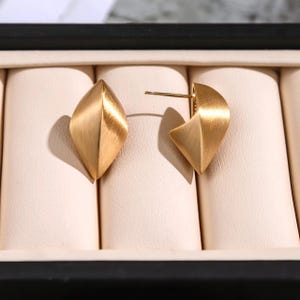 May include: A pair of gold-colored stud earrings. Each earring has a geometric, leaf-like shape with a brushed metal finish. The earrings are displayed on a cream-colored surface within a black jewelry box.