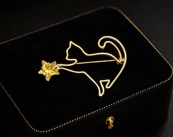 May include: A gold-toned metal brooch shaped like a cat silhouette with a curved tail. The cat is reaching towards a faceted, yellow star-shaped crystal attached to its front paw. The brooch features a thin wire frame design and a pin back mechanism.