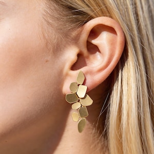 May include: Gold-coloured statement earrings with an abstract design of layered geometric shapes. The earrings are made up of various flat, irregular forms, creating a cluster that dangles from the ear. The earrings are shown against a background of blonde hair and skin.