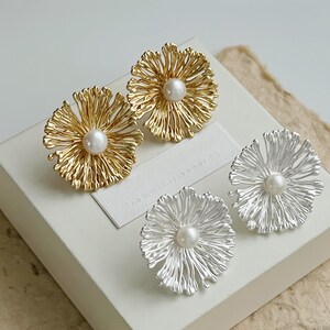 May include: Two pairs of flower-shaped earrings, one pair in gold tone and one pair in silver tone. Each earring features a textured, radiating petal design with a central white pearl. A small white rectangular tag with the text the minority is amazing.
