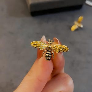 May include: A gold-colored bee brooch with yellow enamel wings and a black and gold striped body. The brooch is held in a person's hand. Another bee brooch is visible in the background.