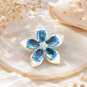 May include: A flower-shaped brooch featuring five pear-cut bright blue gemstones arranged as petals, alternating with five smooth white shell-like petals. The center of the flower is accented with a cluster of small, clear, round-cut stones.