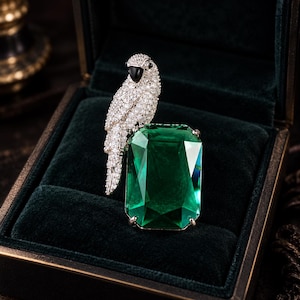May include: A silver-toned ring featuring a large, rectangular, emerald-green faceted stone and a parrot-shaped accent encrusted with numerous small, clear, sparkling stones. The parrot has a smooth, black, teardrop-shaped beak detail.