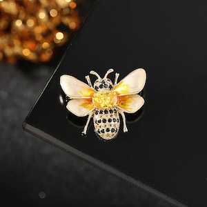 Puede incluir: Broche de abeja dorada con alas color crema, centro amarillo y cuerpo negro y dorado. El broche está adornado con pequeñas piedras preciosas negras y amarillas. La abeja está sobre un fondo negro.