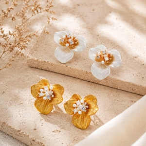Op de afbeelding: Twee paar bloemvormige oorknopjes. Eén paar is zilverkleurig met goudkleurige centra, en het andere paar is goudkleurig. Beide hebben gestructureerde bloemblaadjes en kleine, heldere, fonkelende steentjes en witte kraaltjes in het midden.