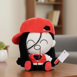 Puede incluir: Muñeco de peluche con gorra de béisbol roja, pelo negro y cara blanca con antifaz negro. El muñeco lleva un chaleco rojo y sostiene una pequeña cuchilla blanca. El muñeco está sentado.