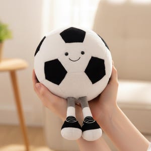 Könnte beinhalten: Ein Plüschfußball mit einem lächelnden Gesicht, schwarz-weißen Paneelen, grauen Beinen und schwarz-weißen Schuhen. Das Stofftier ist als Fußball gestaltet.