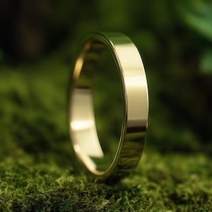 May include: A gold wedding ring with a flat design. The ring is displayed on a bed of green moss, giving a natural and refined look. The polished surface of the band reflects light, emphasising its smooth, clean lines.