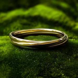 May include: A gold bangle bracelet with three interwoven bands sits on green moss. The polished surface of the bracelet reflects light. The jewelry is a simple, elegant accessory. The bracelet is a stylish piece of jewellery.
